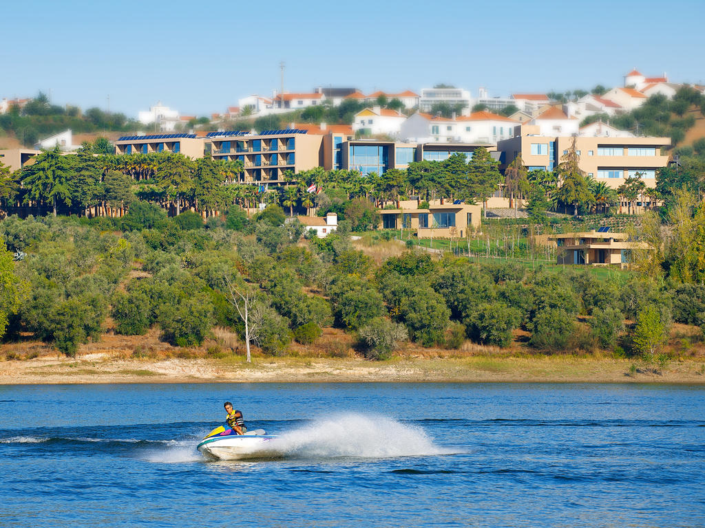 Ap Lago Montargil - Conference & Spa Eksteriør billede The photo depicts a person riding a jet ski on a body of water. In the background, there are modern buildings and lush greenery, suggesting a scenic landscape, likely near a resort or upscale residential area. The clear blue sky and calm water enhanc