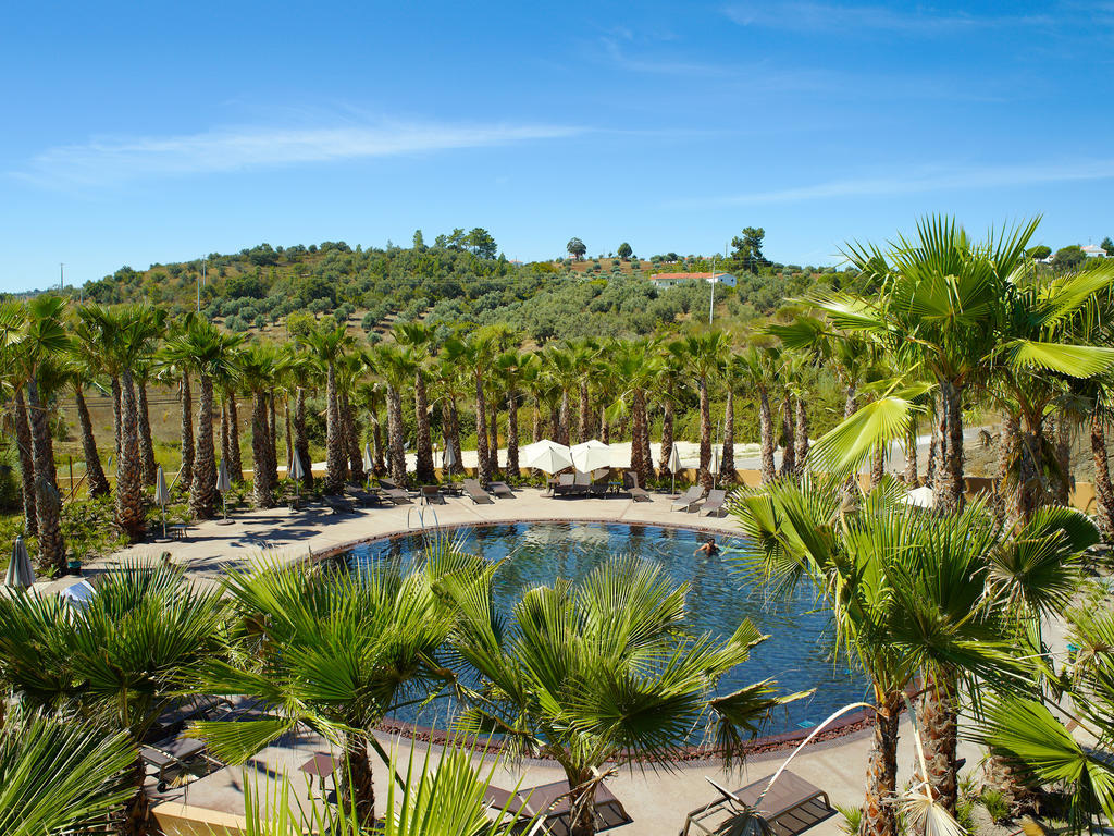 Ap Lago Montargil - Conference & Spa Eksteriør billede The photo depicts a beautiful outdoor area featuring a circular swimming pool surrounded by palm trees. The pool area has several lounge chairs arranged around it. In the background, there is a lush green hillside, and the sky is clear with a bright 