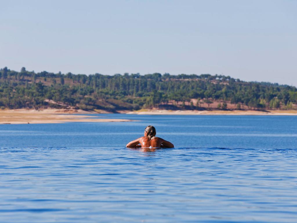 Ap Lago Montargil - Conference & Spa Eksteriør billede The photo shows a person swimming in a calm body of water, likely a lake or a river. The swimmer is seen from behind and appears to be enjoying the water on a sunny day. In the background, there's a scenic landscape featuring green trees and a sandy 