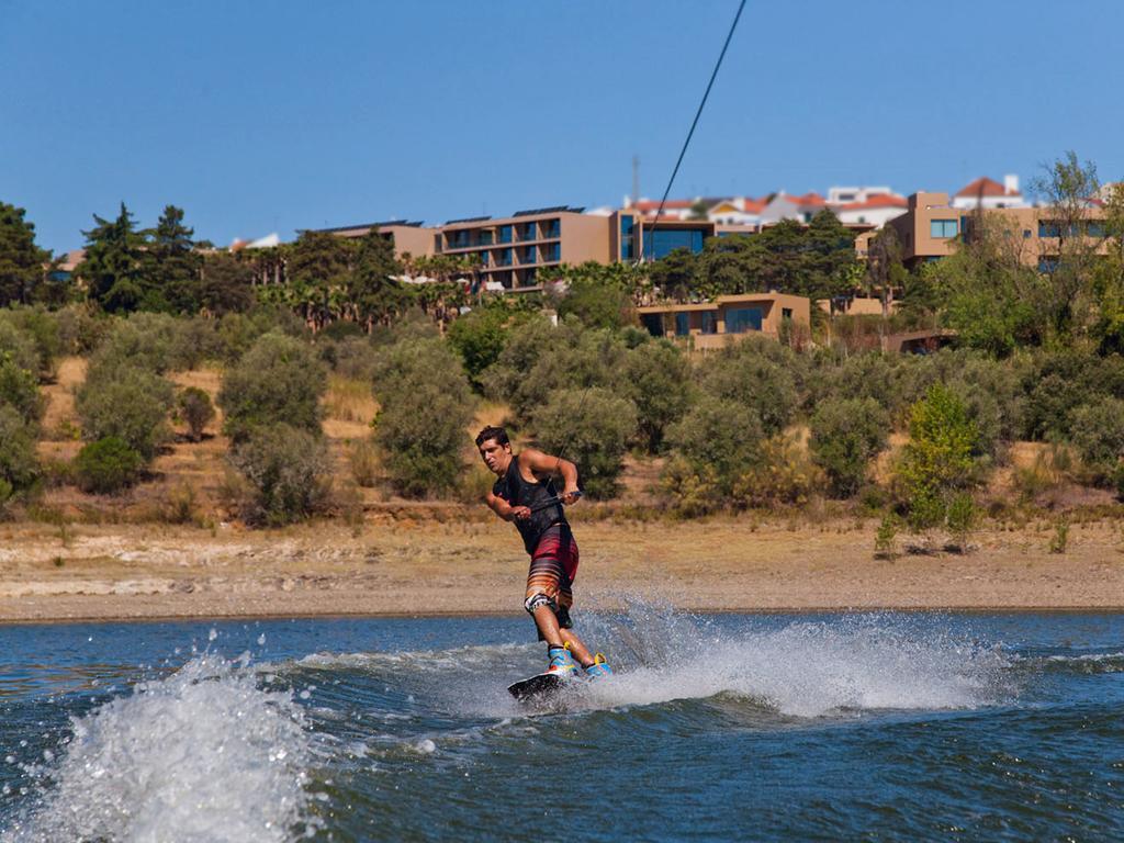 Ap Lago Montargil - Conference & Spa Eksteriør billede The photo shows a person wakeboarding on a body of water. The individual is being pulled by a cable, which is likely connected to a boat or a winch. The person is wearing a life jacket and swim shorts, balancing on the board as they enjoy the activit