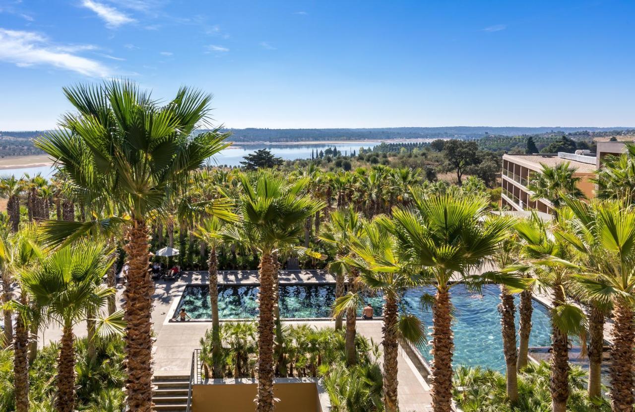 Ap Lago Montargil - Conference & Spa Eksteriør billede The photo depicts a picturesque resort landscape with a clear blue sky. In the foreground, there are tall palm trees surrounding a swimming pool area. The swimming pool has a modern design and is surrounded by lush greenery. In the background, you ca