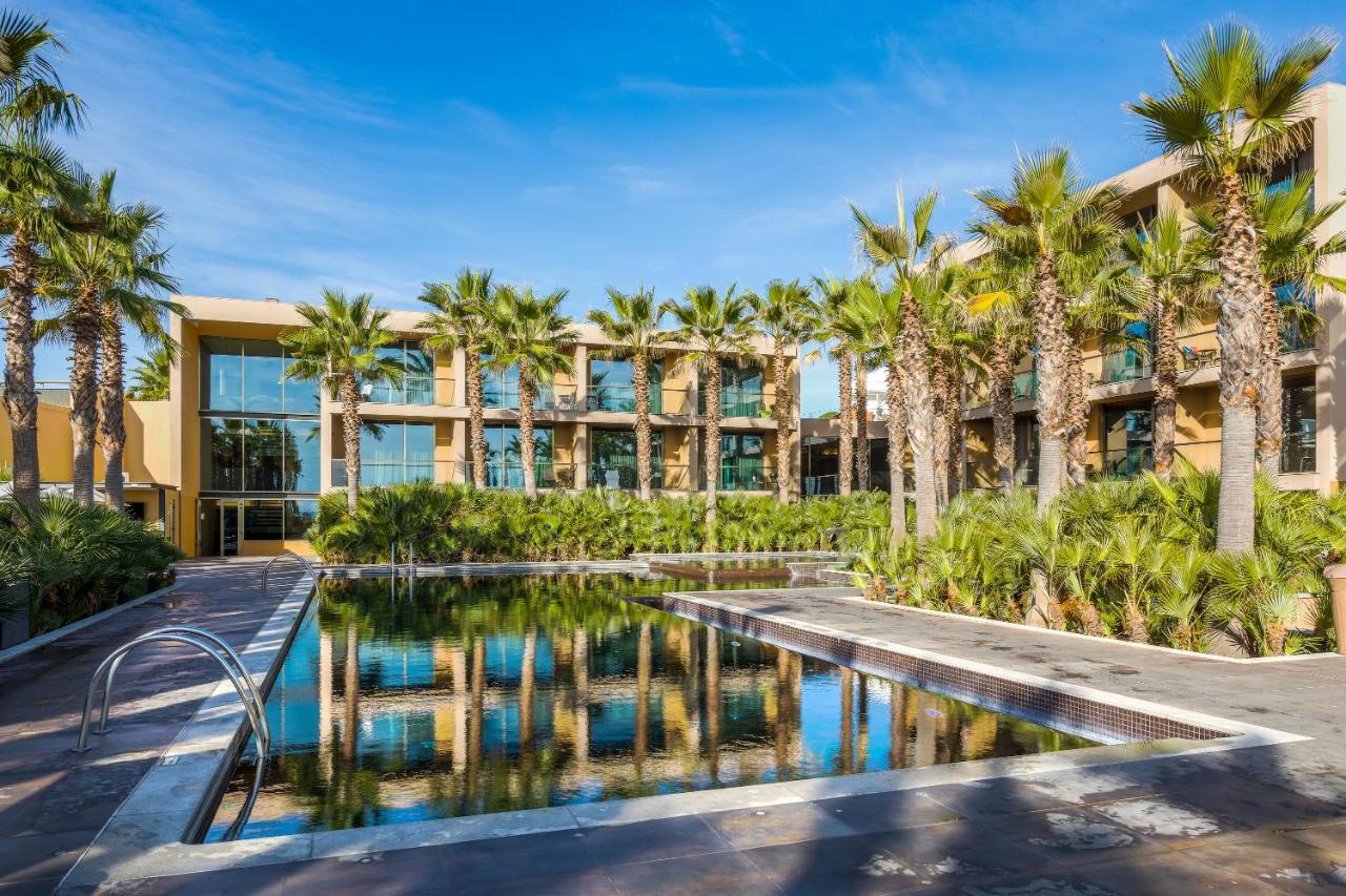 Ap Lago Montargil - Conference & Spa Eksteriør billede The photo features a modern building surrounded by lush greenery and palm trees. In the foreground, there is a serene swimming pool with a reflective surface that captures the surrounding environment. The architecture of the building appears to have 