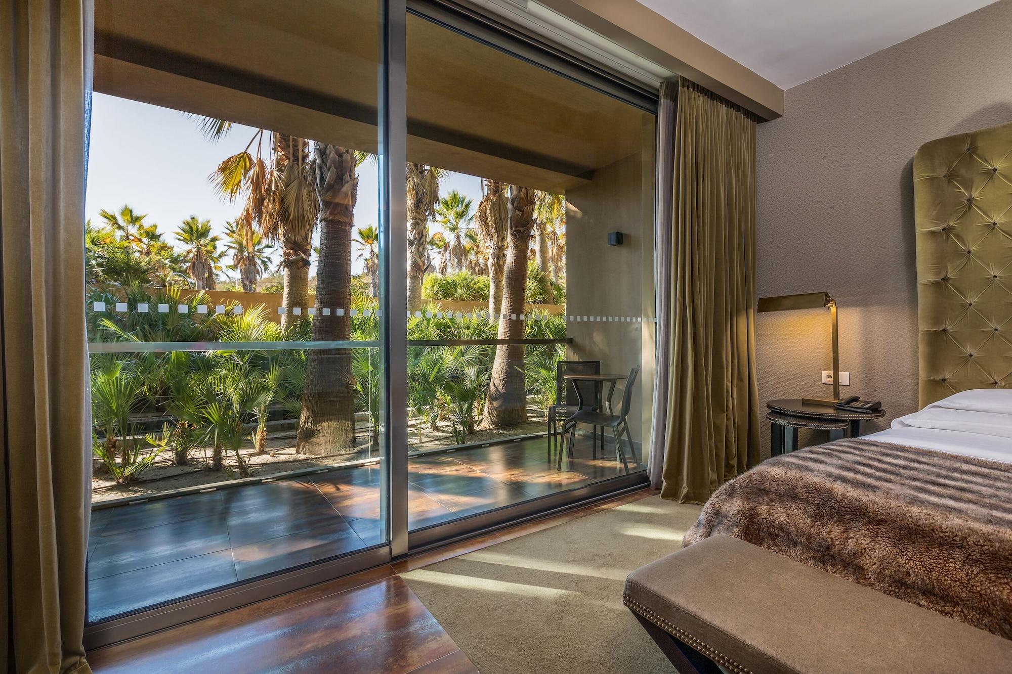 Ap Lago Montargil - Conference & Spa Eksteriør billede The photo shows a bedroom with large glass doors that lead out to a balcony or terrace. The view outside features palm trees and lush greenery, indicating a tropical or warm climate. Inside the room, there is a bed, a bedside table with a lamp, and a