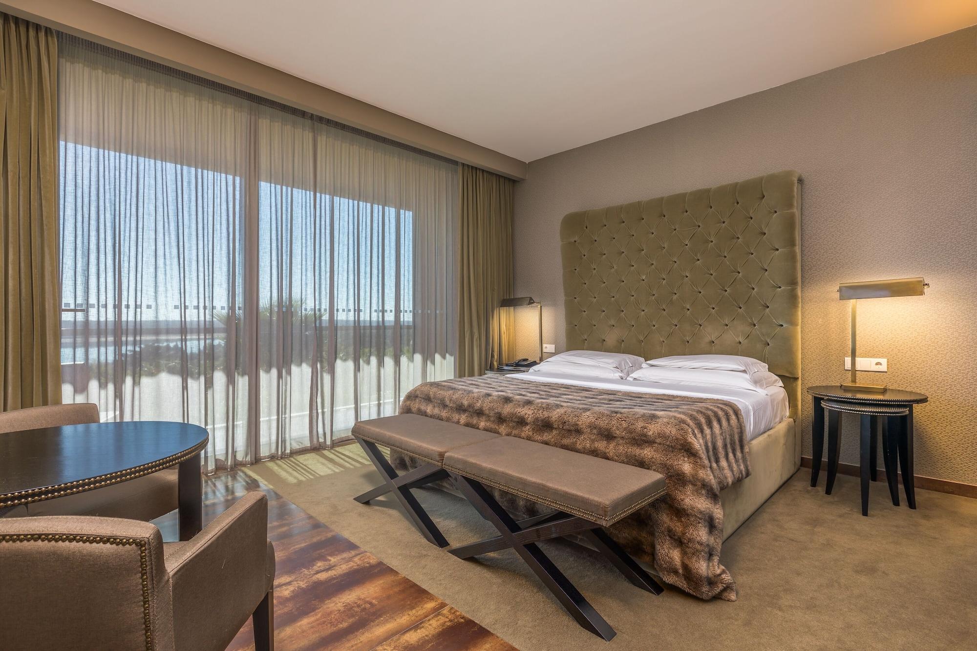 Ap Lago Montargil - Conference & Spa Eksteriør billede The photo shows a modern hotel room featuring a large bed with a tufted headboard and a cozy blanket. There are curtains draped over large windows that allow natural light to flow in, providing a view of the outside. A small seating area includes two