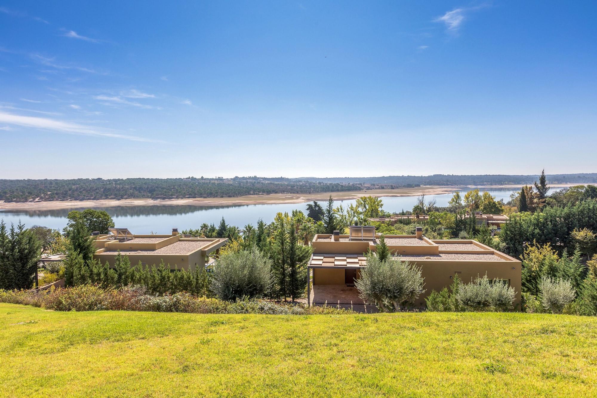 Ap Lago Montargil - Conference & Spa Eksteriør billede The photo depicts a scenic landscape featuring a calm body of water, possibly a lake or river, surrounded by lush greenery and trees. In the foreground, there are several modern buildings with flat roofs, set against a vibrant blue sky. The overall s