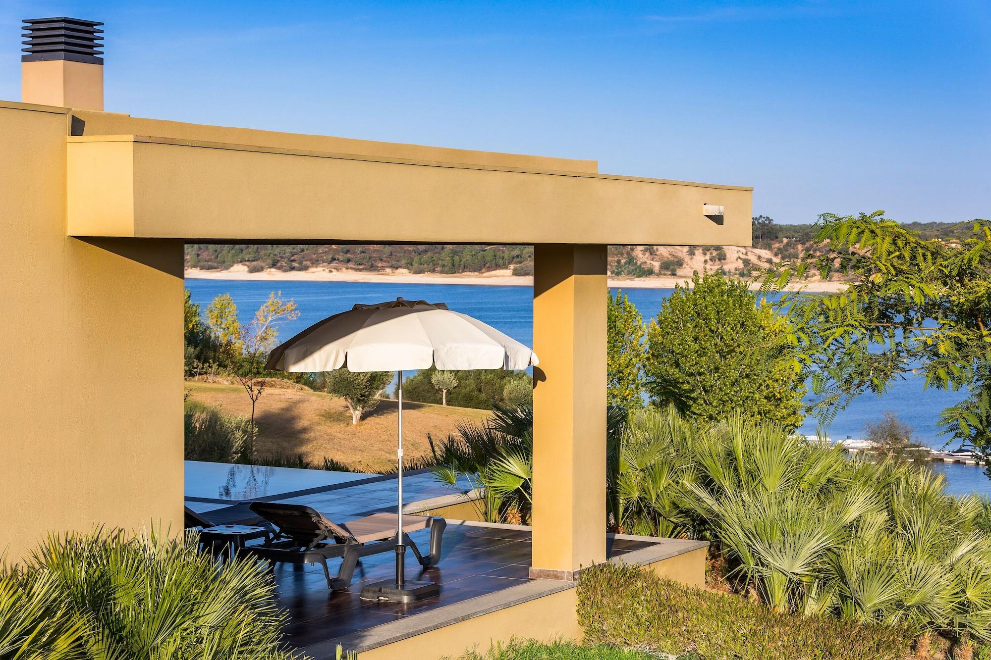 Ap Lago Montargil - Conference & Spa Eksteriør billede The photo shows a peaceful outdoor area with a modern architectural structure featuring an overhang. There is a lounge chair positioned beneath a large umbrella, creating a shaded spot to relax. In the background, there is a picturesque view of a bod