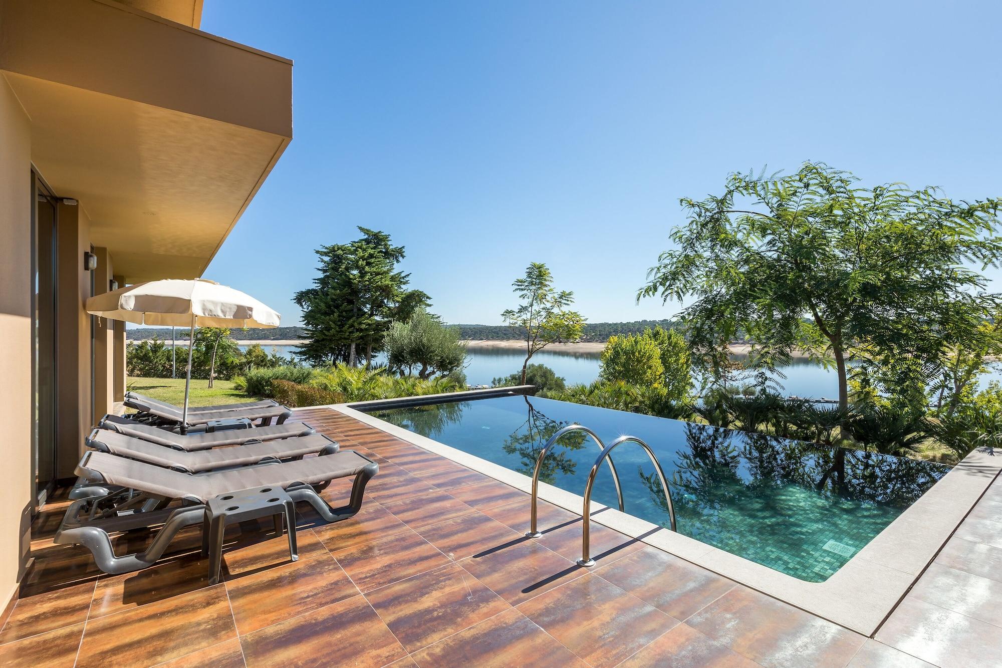 Ap Lago Montargil - Conference & Spa Eksteriør billede The photo shows a luxurious outdoor setting by a pool. There are several lounge chairs arranged next to an infinity pool, which overlooks a scenic body of water and lush greenery. A parasol provides shade for relaxation. The sky is clear and blue, in