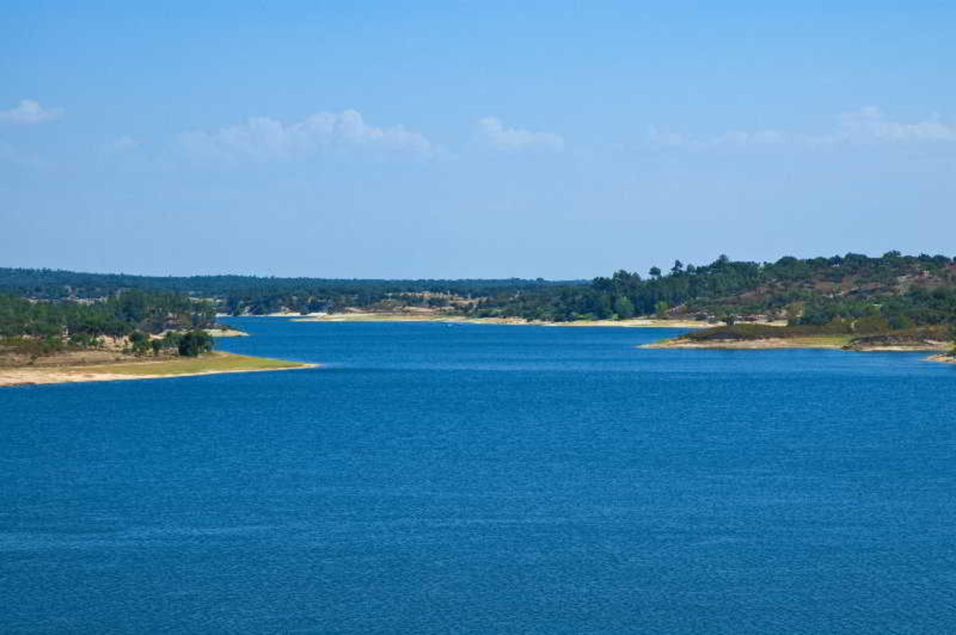 Ap Lago Montargil - Conference & Spa Eksteriør billede The photo depicts a serene landscape featuring a large body of water, likely a lake or river. The water appears calm and is reflecting a clear blue sky with some clouds. Surrounding the water, there are patches of green land and trees, indicating a n