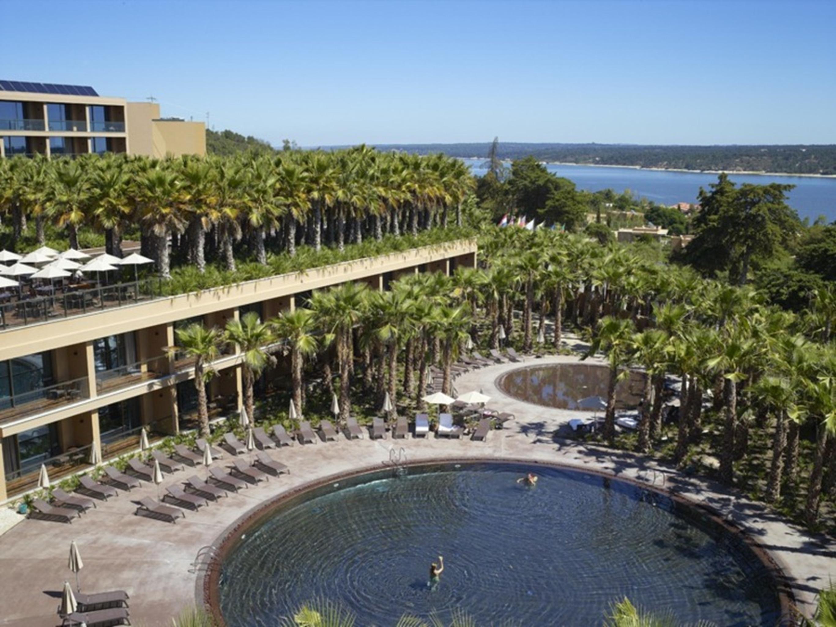 Ap Lago Montargil - Conference & Spa Eksteriør billede The photo shows a scenic view of a resort area. In the foreground, there are circular pools surrounded by lounge chairs, with a few people enjoying the water. Palm trees line the edges, creating a tropical atmosphere. In the background, there is a mu