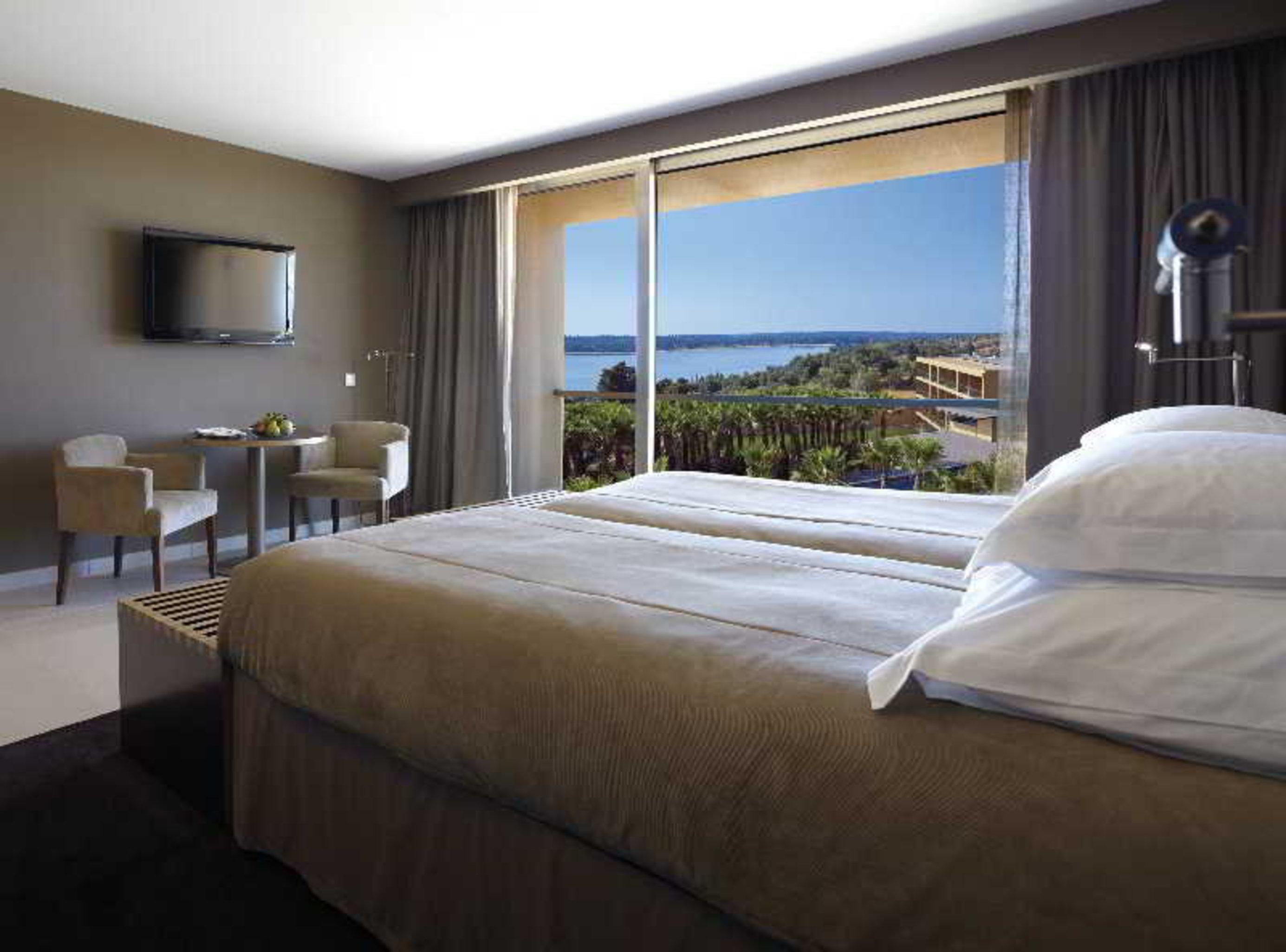 Ap Lago Montargil - Conference & Spa Eksteriør billede The photo shows a modern hotel room with a large window offering a view of the sea and mountains. The room features a comfortable bed with white pillows and brown bedding. There is a small sitting area with two chairs and a table, as well as a wall-m