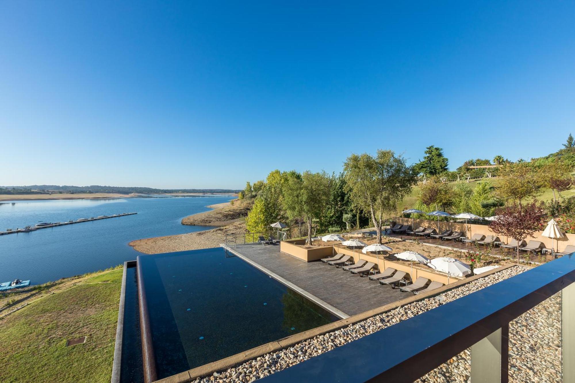 Ap Lago Montargil - Conference & Spa Eksteriør billede The photograph depicts a scenic view of a tranquil water body, likely a lake or river, bordered by lush greenery. In the foreground, there is an infinity pool that blends seamlessly with the water beyond. Comfortable lounge chairs are arranged on a w