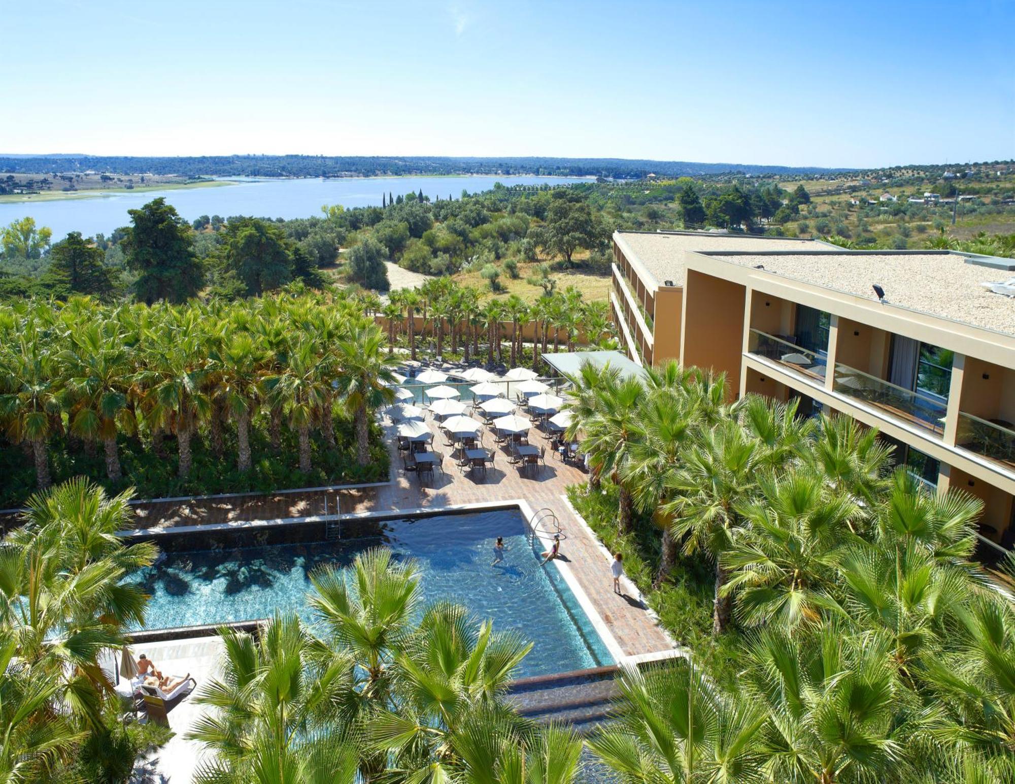 Ap Lago Montargil - Conference & Spa Eksteriør billede The photo shows a scenic view of a modern hotel or resort. In the foreground, there is a swimming pool surrounded by palm trees, with lounge chairs positioned beside it. Sun umbrellas provide shade over the chairs. The background features a lush land